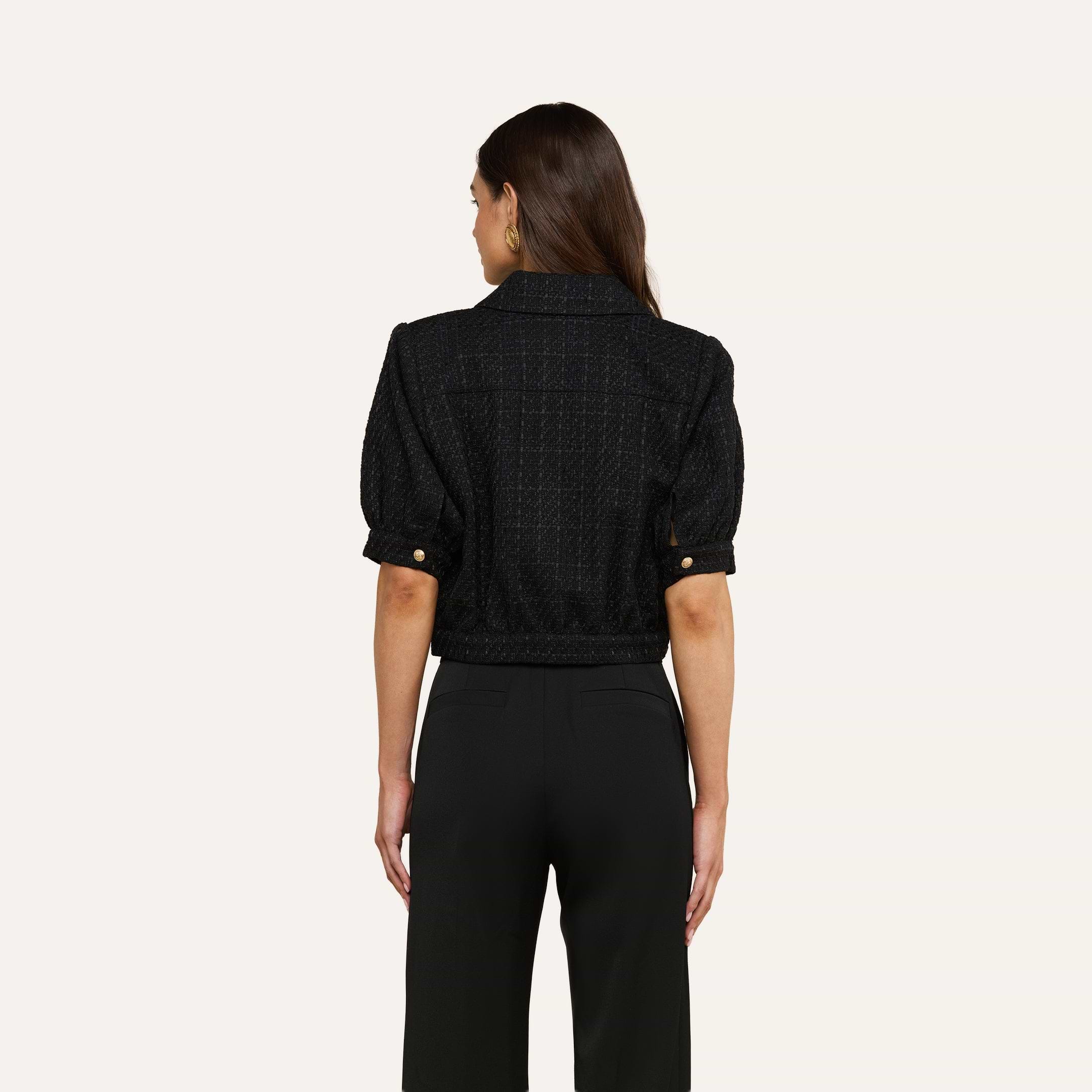 A woman with long brown hair stands facing away from the camera against a plain light background, wearing black pants and the Cove Cropped Tweed Jacket featuring a cropped silhouette.