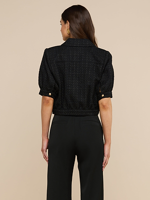 Eine Frau mit langen braunen Haaren ist von hinten zu sehen. Sie trägt die „Cove Cropped Tweed Jacket“ mit goldenen Knöpfen und eine schwarze Hose mit hoher Taille und steht vor einem beigen Hintergrund. Die verkürzte Silhouette der Jacke verleiht dem Outfit einen modernen Touch.