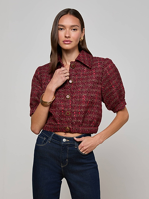 A woman with long brown hair wears the L'AGENCE Cove Cropped Tweed Jacket, featuring vintage-inspired buttons and short sleeves, paired with dark blue high-waisted jeans. She stands with one hand on her waist against a plain light background.