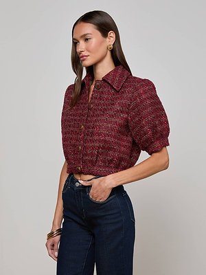 A woman with long brown hair wears the L'AGENCE Cove Cropped Tweed Jacket, styled with dark blue jeans and gold accessories, standing against a plain light background.