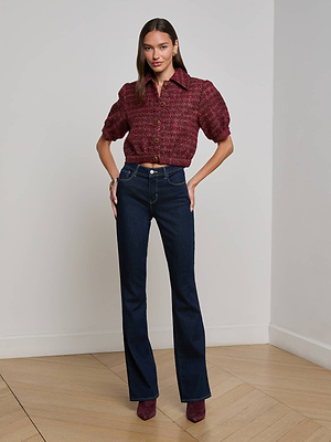 A woman stands indoors on wooden flooring, facing forward with hands on hips, wearing the L'AGENCE Cove Cropped Tweed Jacket, dark high-waisted jeans, and burgundy pointed-toe shoes.