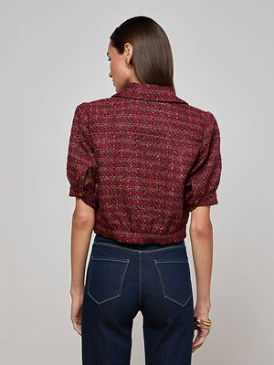 A woman with straight brown hair faces away, wearing the L'AGENCE Cove Cropped Tweed Jacket in textured dark red and high-waisted dark blue jeans, with a gold bracelet on her wrist, set against a plain light gray background.