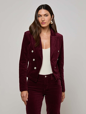 A woman models the L'AGENCE Wayne Velvet Blazer in deep maroon with silver buttons and matching pants, layered over a white top, standing against a plain light gray background.