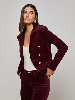 A woman with long brown hair wears the Wayne Velvet Blazer by L'AGENCE in maroon, paired with matching pants and a white top. She stands against a plain light gray background, looking to the side.