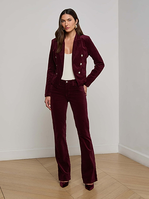A woman poses confidently indoors in the Wayne Velvet Blazer by L'AGENCE, styled with matching flared pants, a white top, and burgundy heels. She has long brown hair and stands with one hand in her pocket.