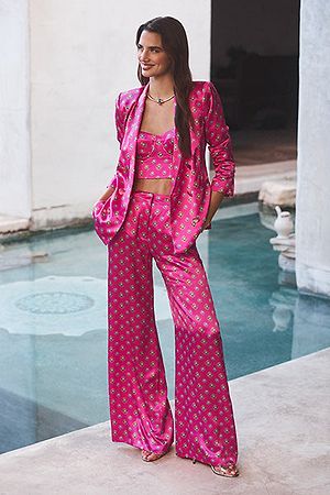 A woman stands by a pool wearing a vibrant pink satin pantsuit with a matching crop top, all featuring a small geometric pattern. She is smiling, with her hands in her pants pockets.