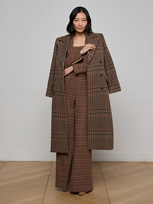 A woman with dark hair poses confidently indoors against a plain light background, wearing the Kolina Plaid Long Coat draped over her shoulders, paired with a tailored blazer and wide-leg trousers.