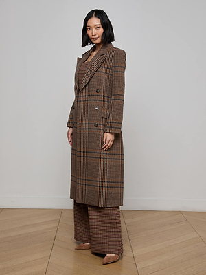 A woman stands indoors against a light wall and wooden floor, wearing the Kolina Plaid Long Coat—a long, brown houndstooth double-breasted coat with matching wide-leg trousers in Italian recycled wool. Her dark hair is shoulder-length and down.