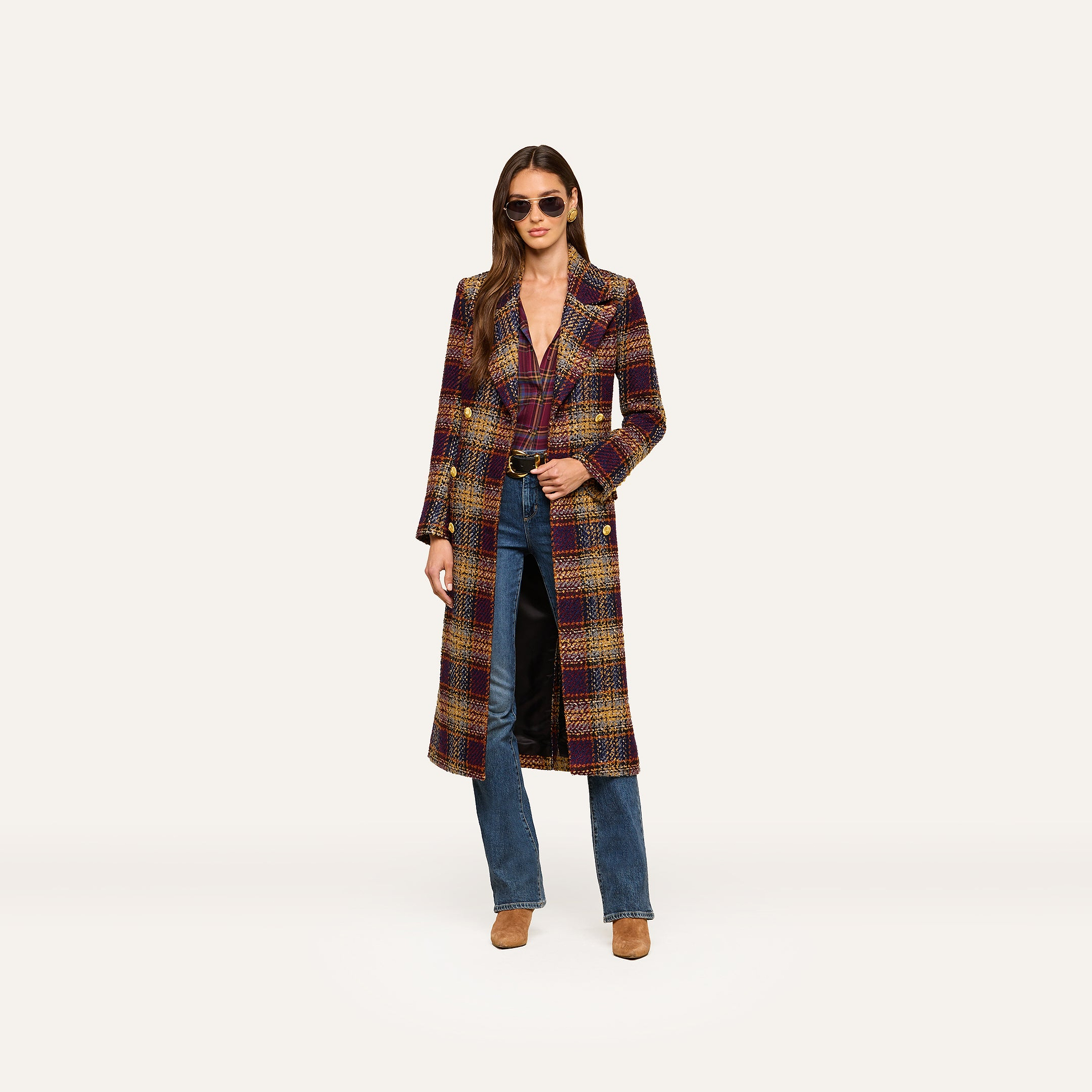A woman wearing sunglasses, the Kolina Tweed Long Coat with a waist-cinching belt, blue jeans, and tan boots stands against a plain white background.