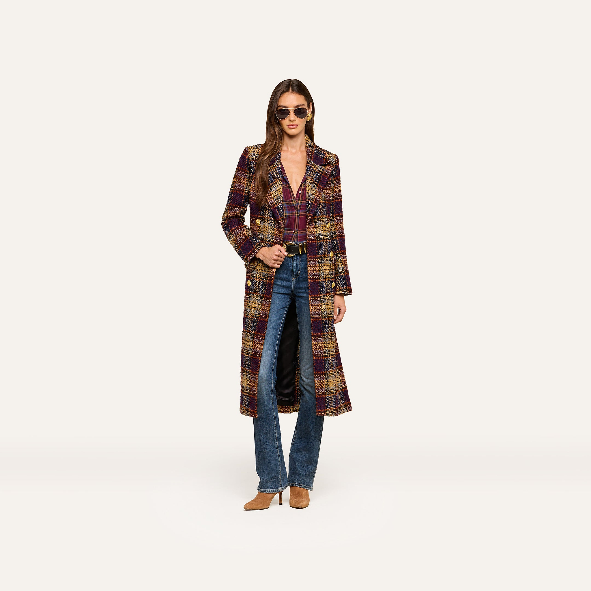 A woman wearing sunglasses, the Kolina Tweed Long Coat with a waist-cinching belt, a purple top, blue jeans, and brown shoes stands against a plain white background.