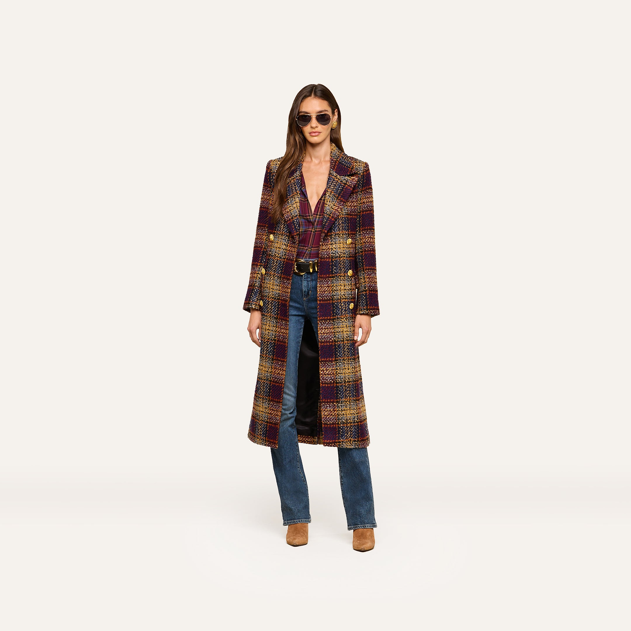 A woman wearing sunglasses and the Kolina Tweed Long Coat with gold buttons, along with blue jeans and brown shoes, stands against a plain light background.