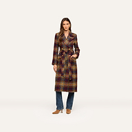 A woman stands against a plain light background wearing the Kolina Tweed Long Coat, which features a waist-cinching belt, styled over blue jeans and tan heeled shoes.