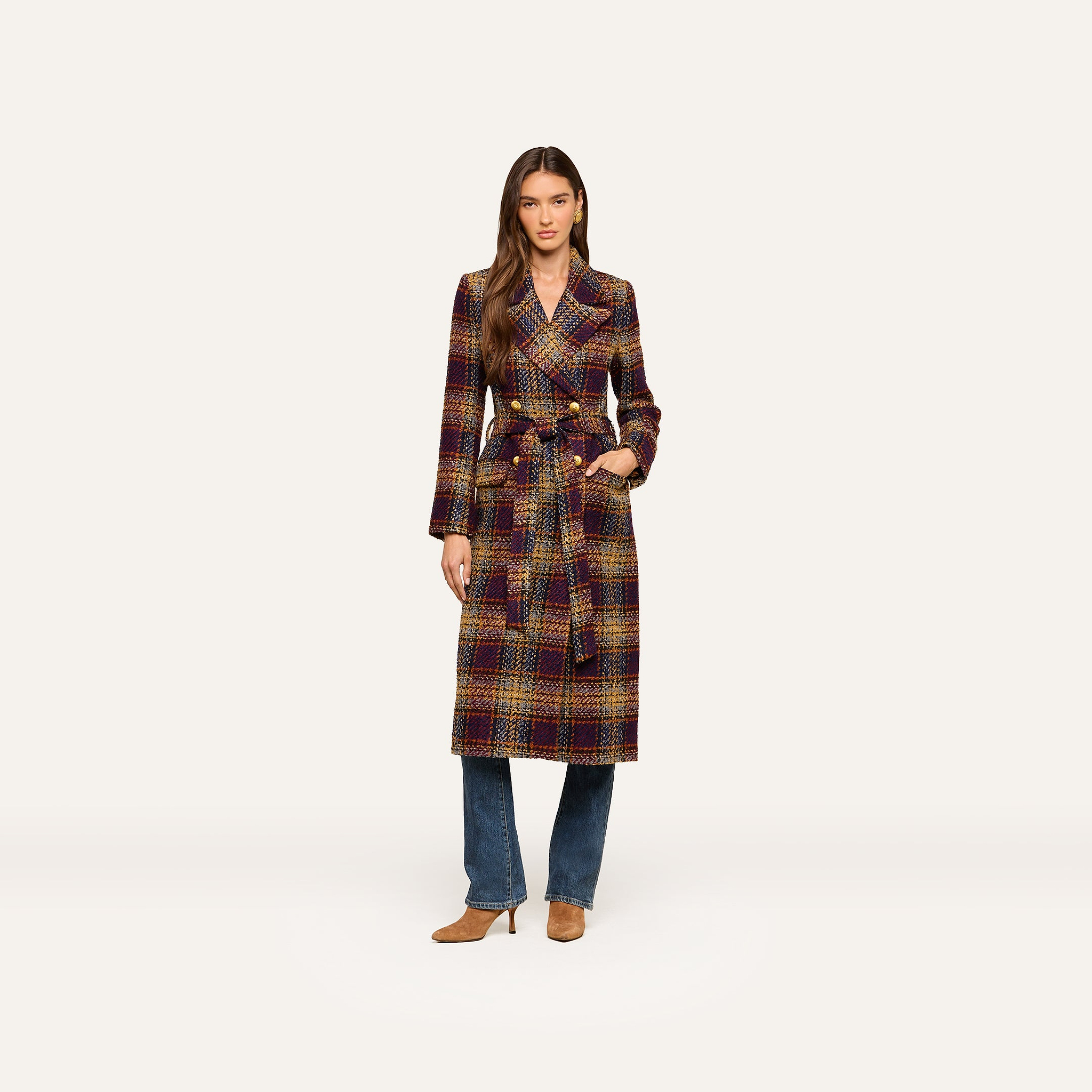 A woman stands against a plain light background wearing the Kolina Tweed Long Coat, which features a waist-cinching belt, styled over blue jeans and tan heeled shoes.