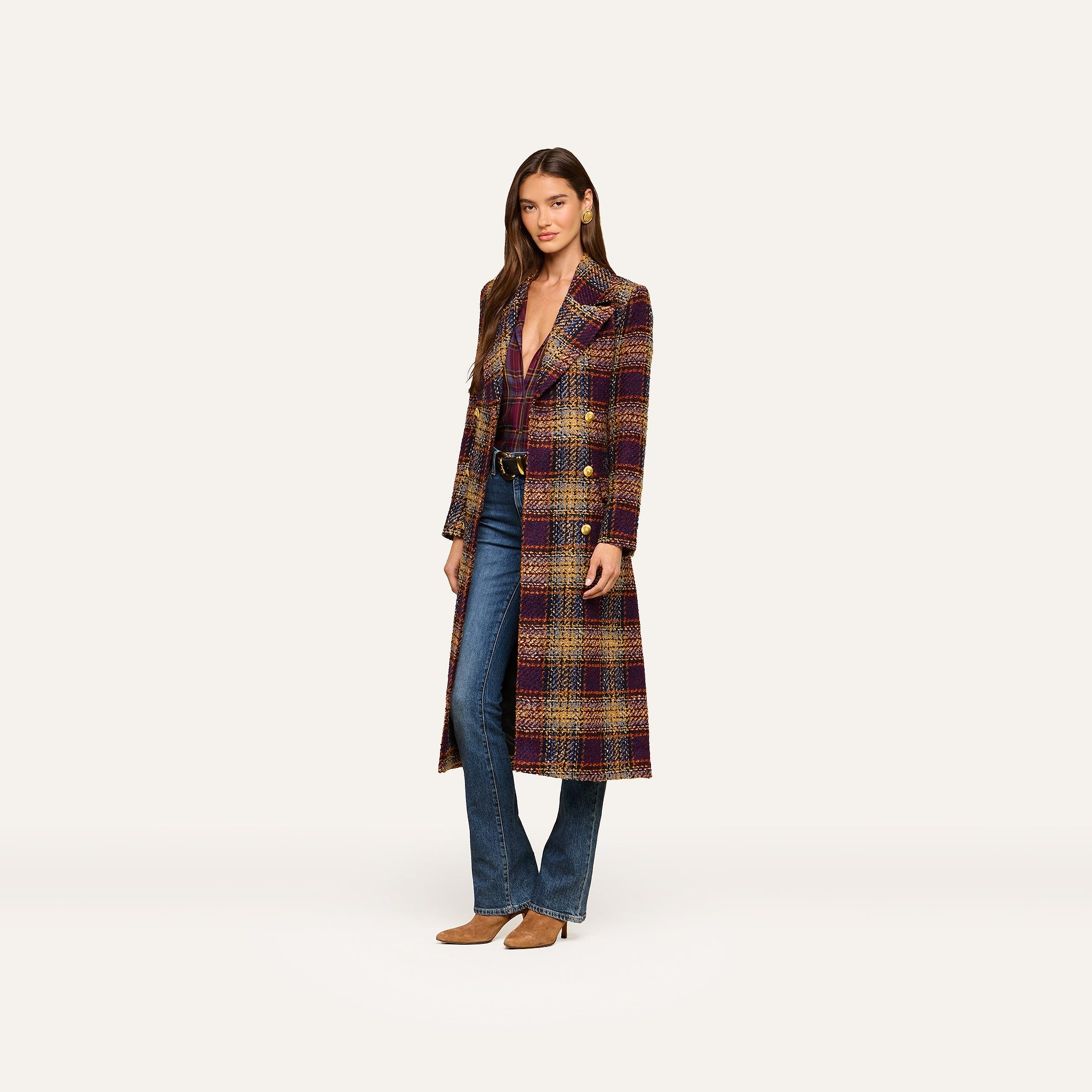 A woman stands against a plain, light background wearing the Kolina Tweed Long Coat—a long, plaid tweed coat with a double-breasted design—layered over a burgundy top, blue jeans, and tan shoes.