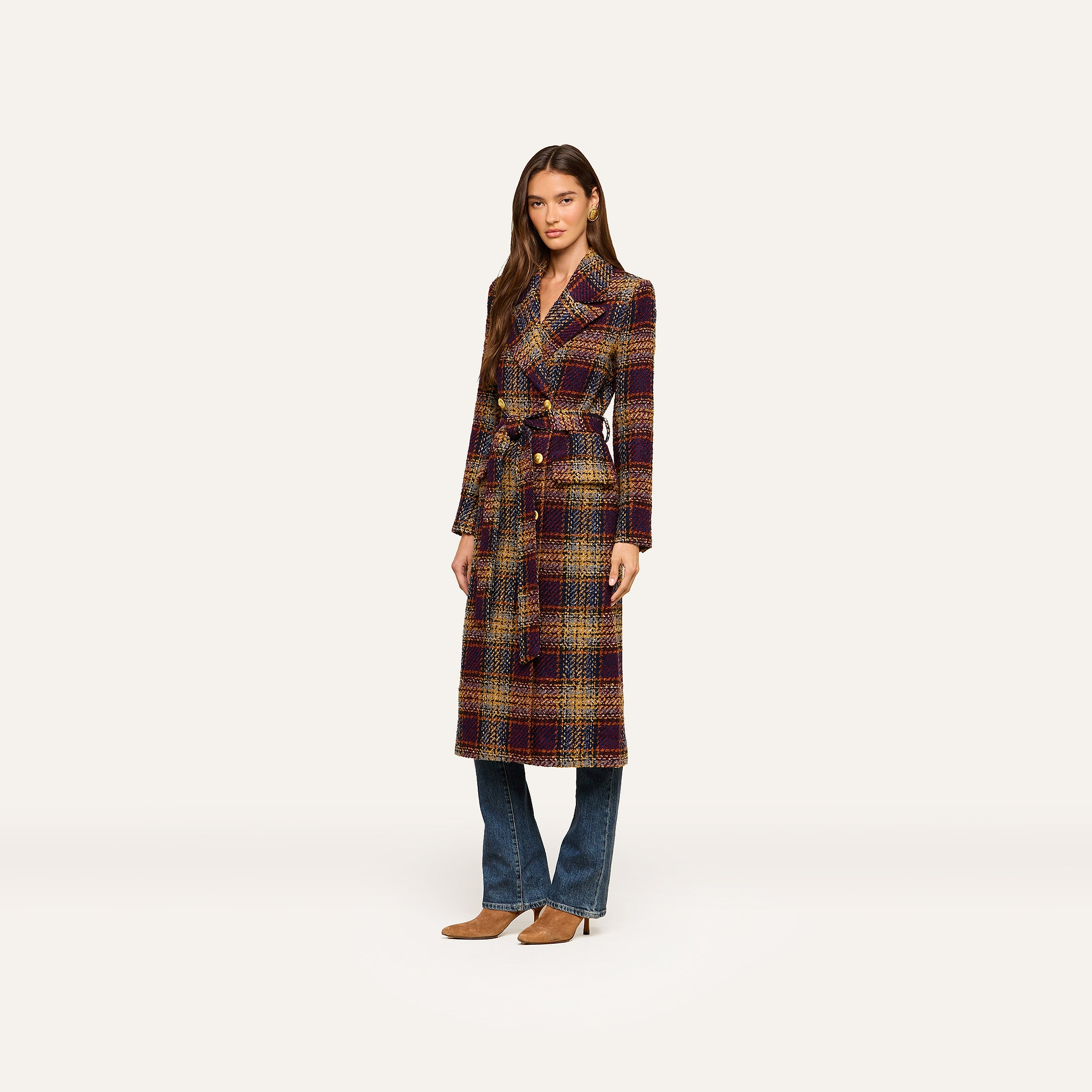 A woman stands against a plain white background wearing the Kolina Tweed Long Coat with a waist-cinching belt, styled over blue jeans and brown shoes.