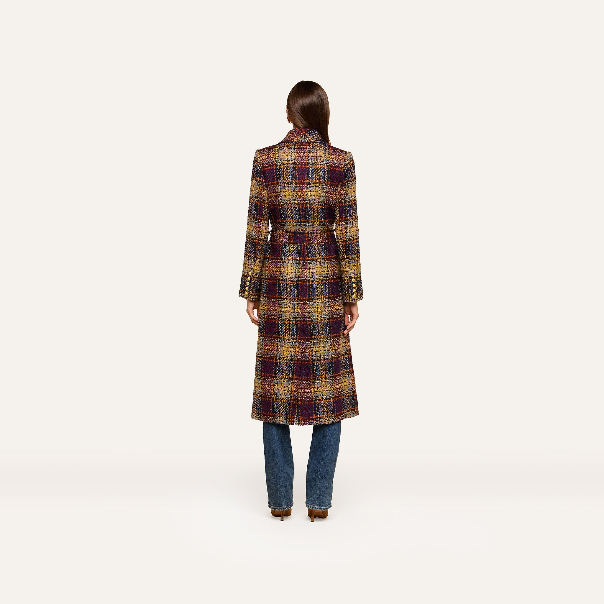 A person stands with their back to the camera, wearing the Kolina Tweed Long Coat—an elegant plaid design featuring a waist-cinching belt—paired with blue jeans against a plain, light background.