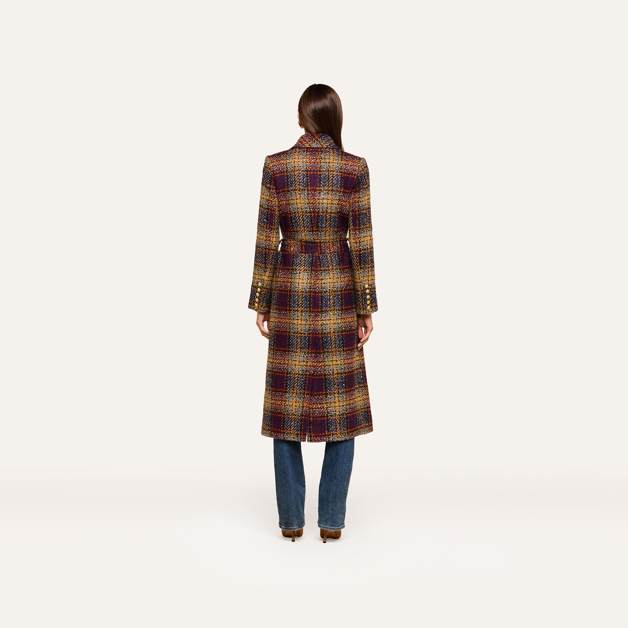A person stands with their back to the camera, wearing the Kolina Tweed Long Coat—an elegant plaid design featuring a waist-cinching belt—paired with blue jeans against a plain, light background.