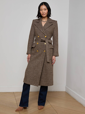 A woman stands indoors wearing the Kolina Long Coat—an elegant, long brown Italian wool coat with gold buttons and a matching belt—paired with dark blue jeans and brown suede shoes, confidently showcasing its sophisticated style.