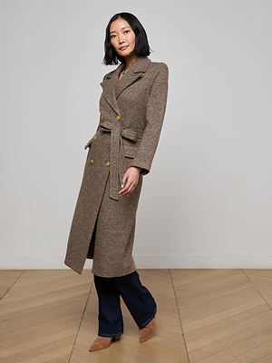A woman poses indoors wearing the Kolina Long Coat with gold buttons, paired with dark blue jeans and tan pointed shoes, standing on a wooden floor against a plain light gray wall.