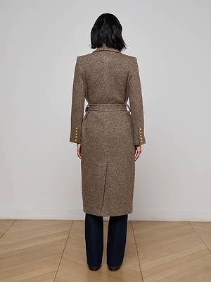 A person faces away wearing the Kolina Long Coat—a long brown tailored coat with a belted waist and gold buttoned sleeves—over dark pants. The setting features a plain white wall and wooden flooring.
