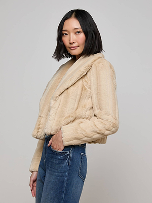 A woman with shoulder-length dark hair models the L'AGENCE Davy Faux Fur Jacket in light beige with blue jeans, standing against a plain light gray background and looking at the camera with a slight smile.