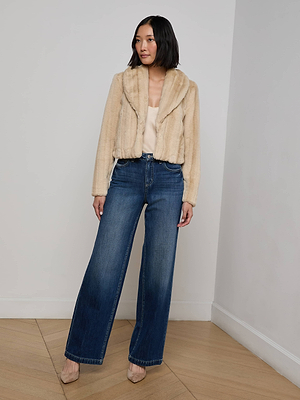 A woman stands indoors against a white wall and light wood floor, wearing the L'AGENCE Davy Faux Fur Jacket in light beige over a cream top, paired with blue wide-leg jeans and beige heels.