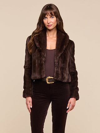 A woman with long brown hair wears the Davy Faux Fur Jacket in dark brown, paired with a brown top and dark pants, standing against a plain beige background.