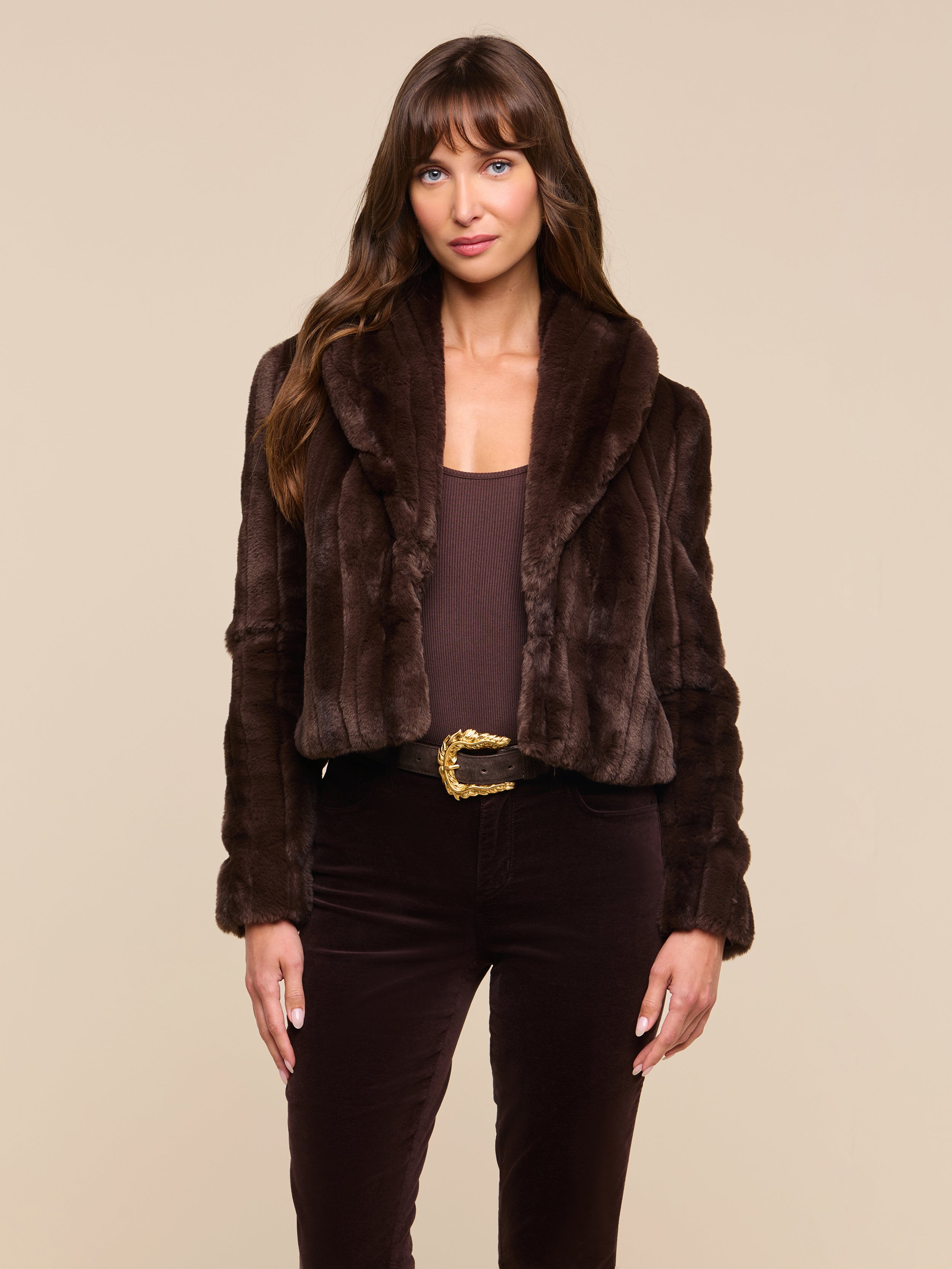 A woman stands facing forward, wearing the Davy Faux Fur Jacket over a brown top and pants with a decorative belt, set against a plain light background.
