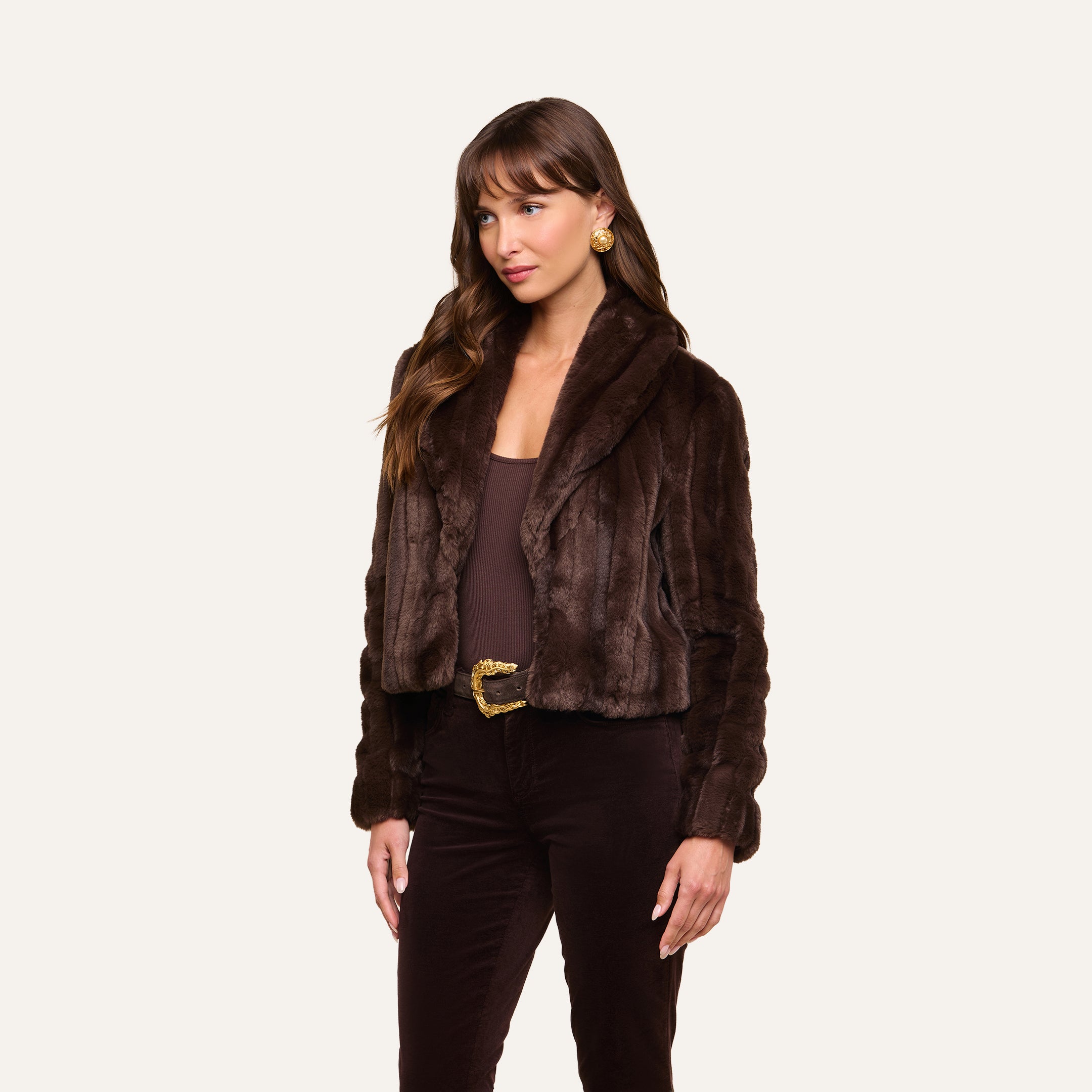 Wearing the Davy Faux Fur Jacket in a retro-inspired dark brown, a woman stands against a plain light background. She pairs the jacket with a brown top, dark pants, a belt, and gold earrings, and her long hair with bangs completes the look.