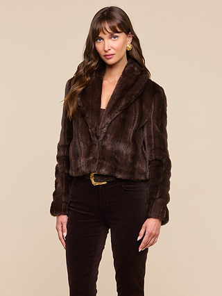 A woman with long brown hair wears the Davy Faux Fur Jacket—cropped and dark brown—paired with gold earrings and dark pants, standing against a beige background and looking at the camera with a relaxed expression.