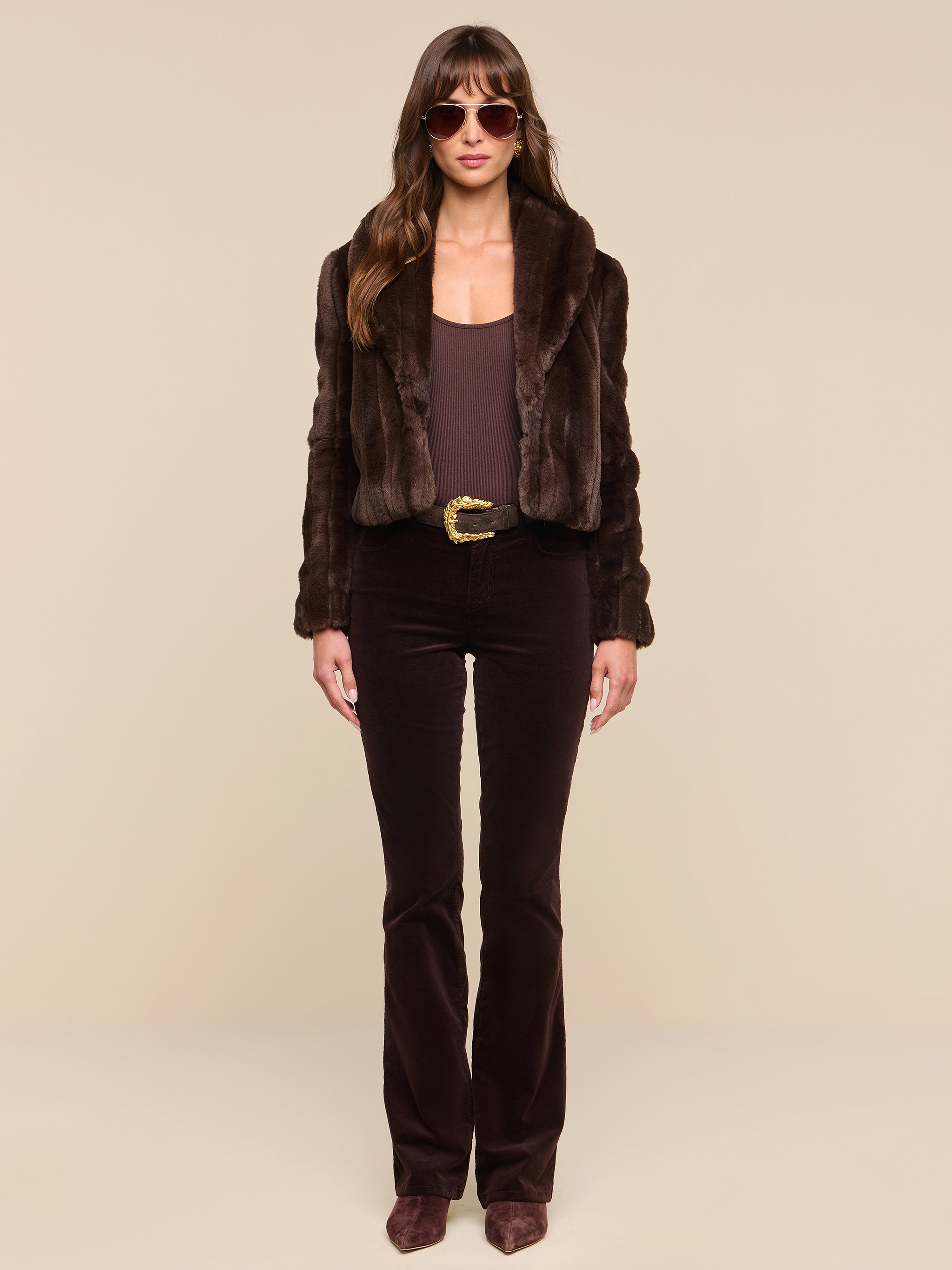 Wearing the Davy Faux Fur Jacket, brown sunglasses, a brown top, a brown belt, dark brown pants, and brown shoes, a woman stands against a plain white background.