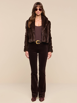 A woman wearing sunglasses, the Davy Faux Fur Jacket, a brown top, flared brown pants, and pointed shoes stands against a plain beige background, facing forward with a neutral expression.