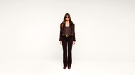 A person with long hair, wearing sunglasses, a dark jacket, dark pants, and brown shoes stands against a plain white background, facing forward.