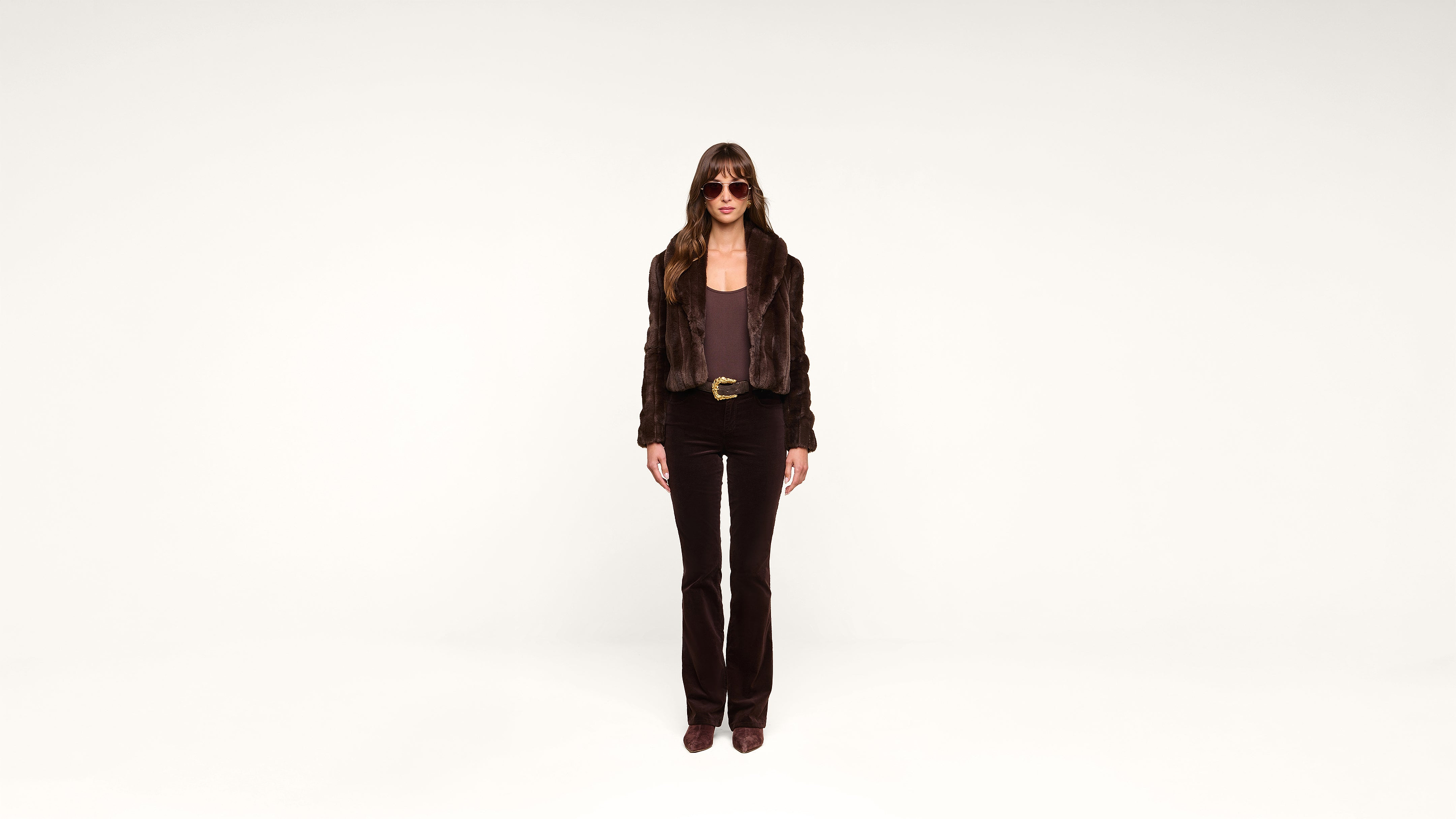 A person with long hair, wearing sunglasses, a dark jacket, dark pants, and brown shoes stands against a plain white background, facing forward.