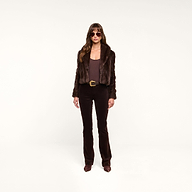 A woman wearing sunglasses, the Davy Faux Fur Jacket, a brown top, brown pants, and pointed brown shoes stands against a plain white background.