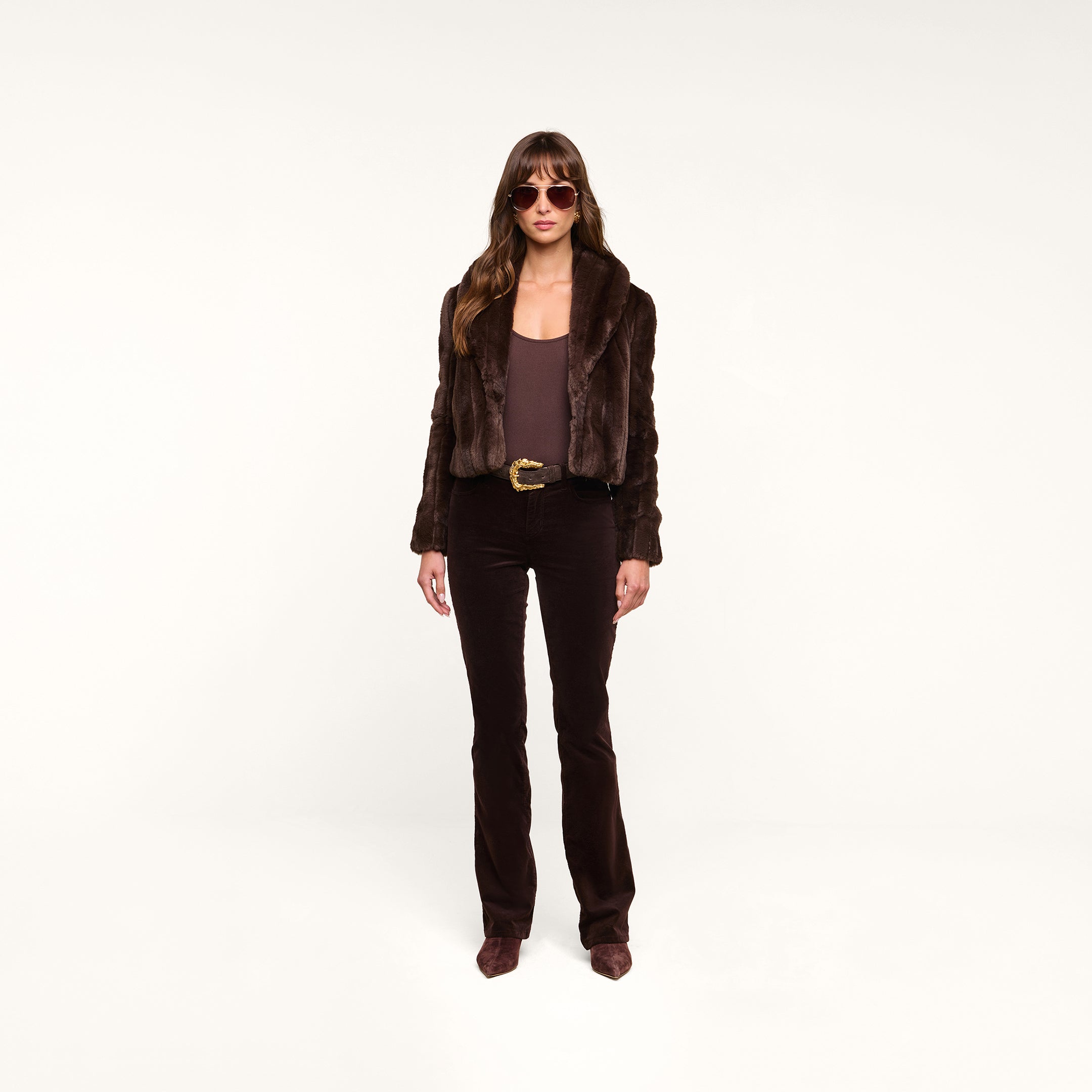 A woman wearing sunglasses, the Davy Faux Fur Jacket, a brown top, brown pants, and pointed brown shoes stands against a plain white background.