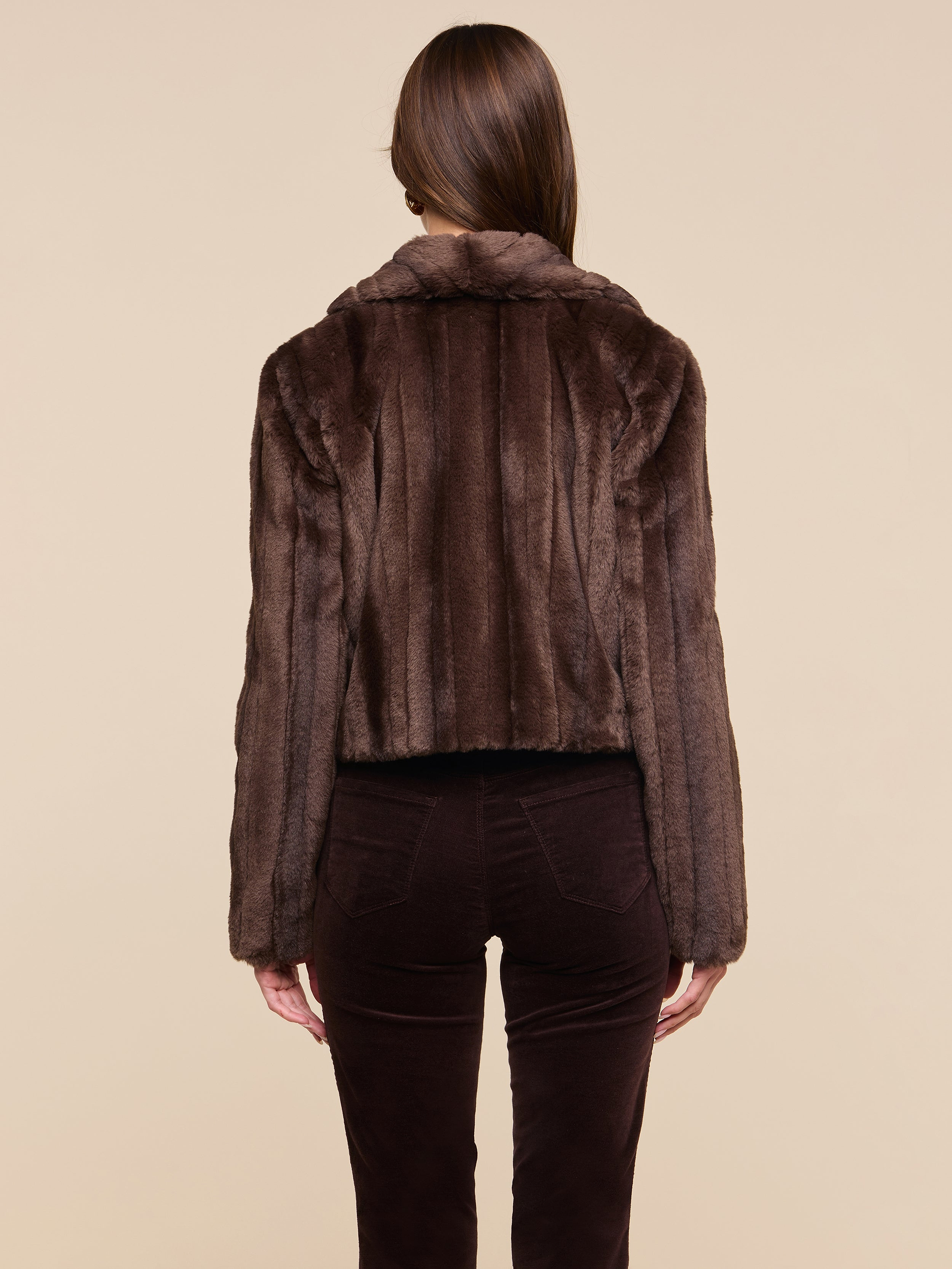 A person with straight brown hair is shown from the back, wearing the retro-inspired Davy Faux Fur Jacket and dark brown pants against a plain light background.