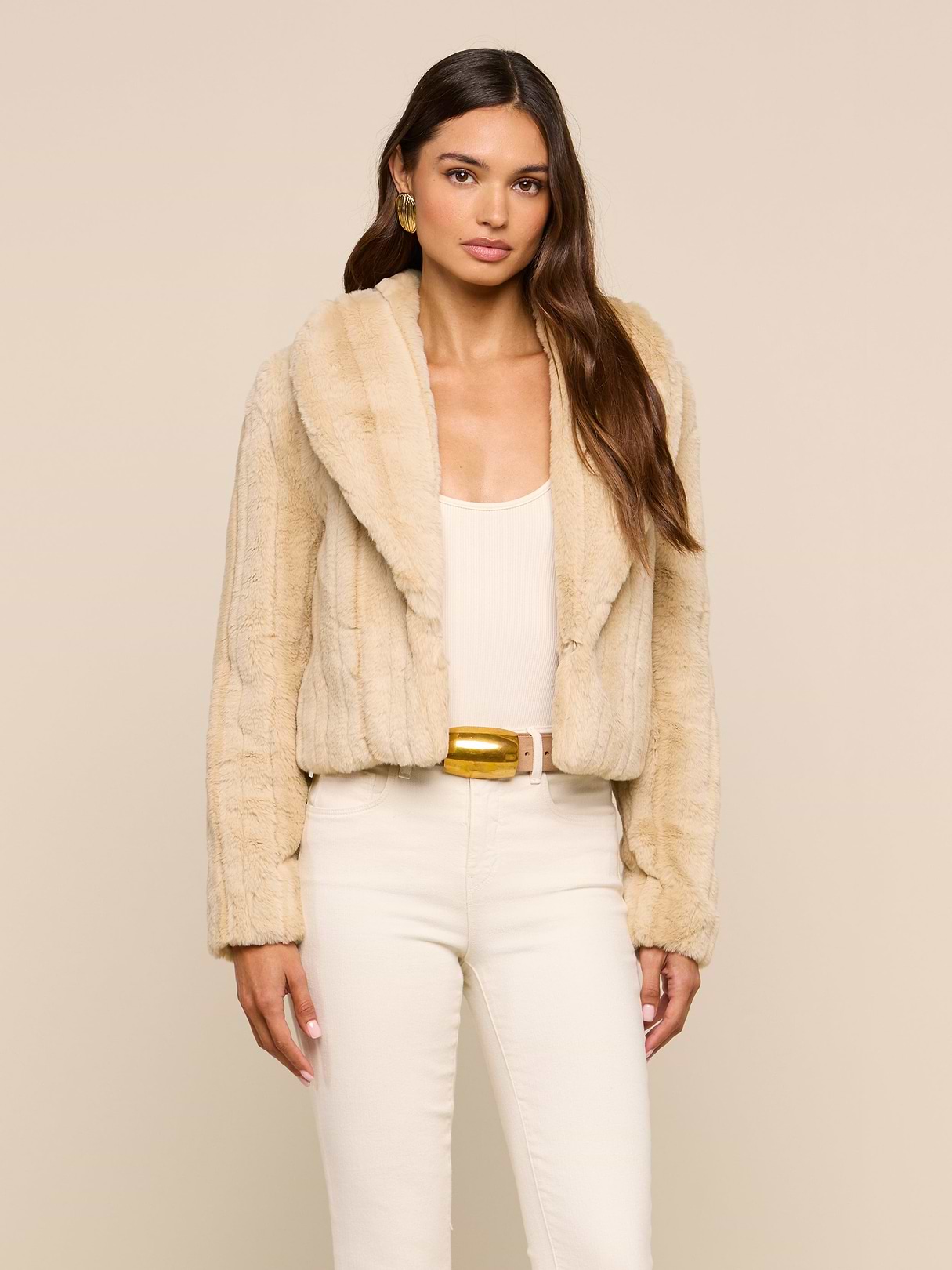 Wearing the Davy Faux Fur Jacket—a beige piece with a subtle mink look—over a cream top and high-waisted pants, a woman stands against a plain beige background, looking at the camera with a neutral expression.