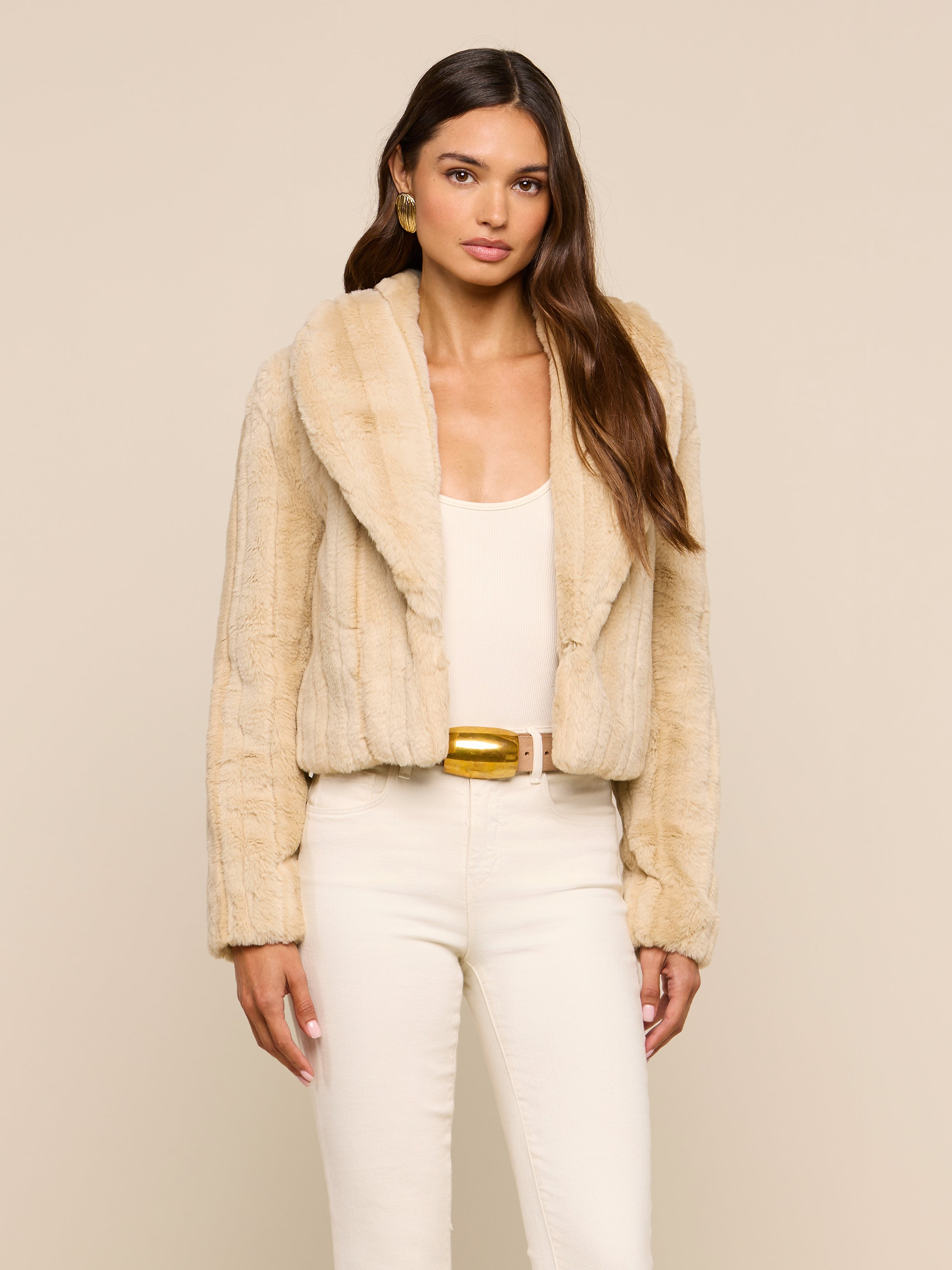 A woman models the Davy Faux Fur Jacket, featuring a luxe beige mink-look finish, styled over a white top and white pants and accessorized with gold earrings and a belt, set against a simple light background.