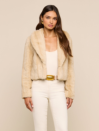 Wearing the Davy Faux Fur Jacket—a beige piece with a subtle mink look—over a cream top and high-waisted pants, a woman stands against a plain beige background, looking at the camera with a neutral expression.