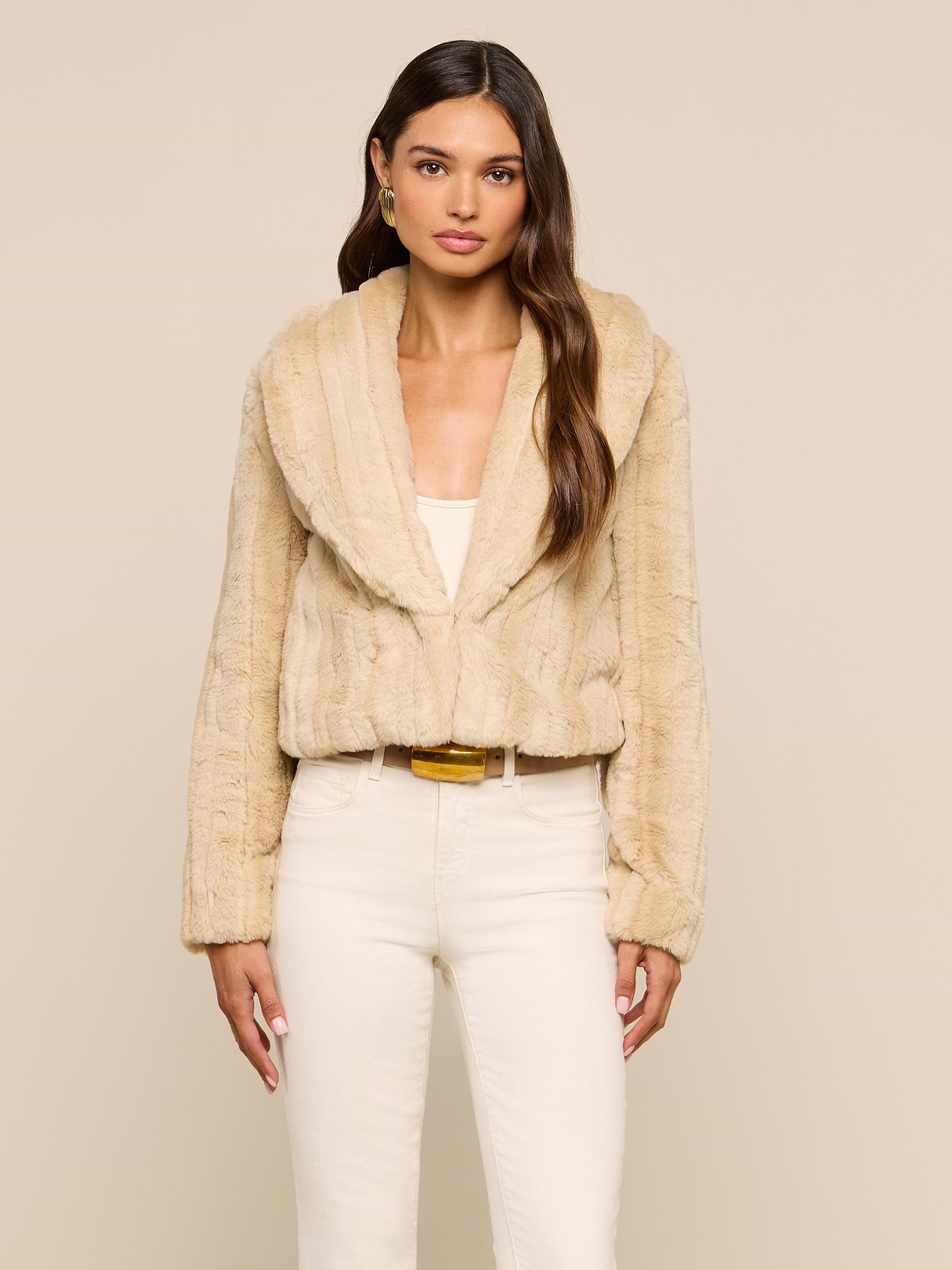 A woman wearing the Davy Faux Fur Jacket in light beige over a white top and cream pants stands against a plain beige background, looking at the camera with a neutral expression.