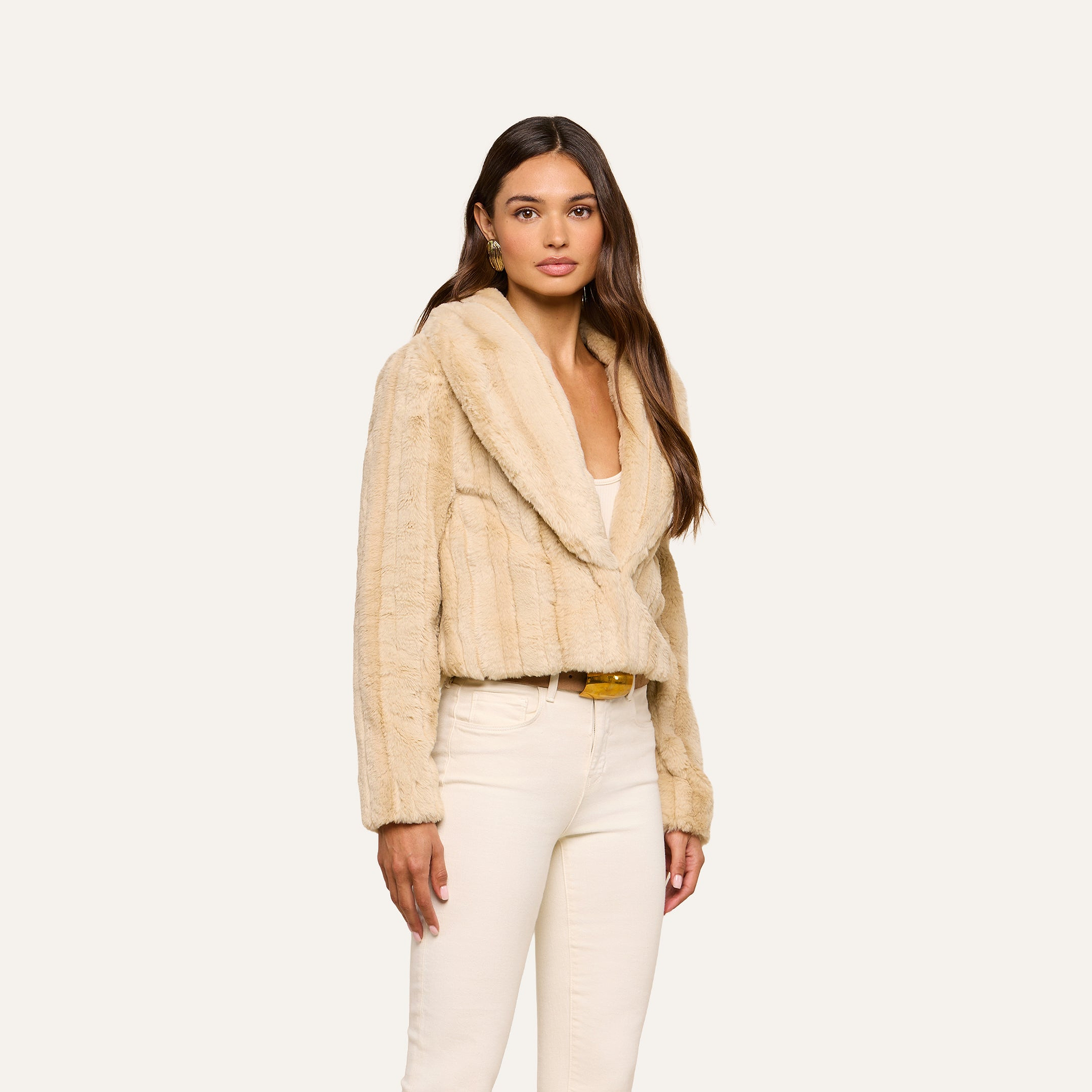 A woman wearing the Davy Faux Fur Jacket with a mink look over a white top and cream pants stands against an off-white background, looking toward the camera with a neutral expression.