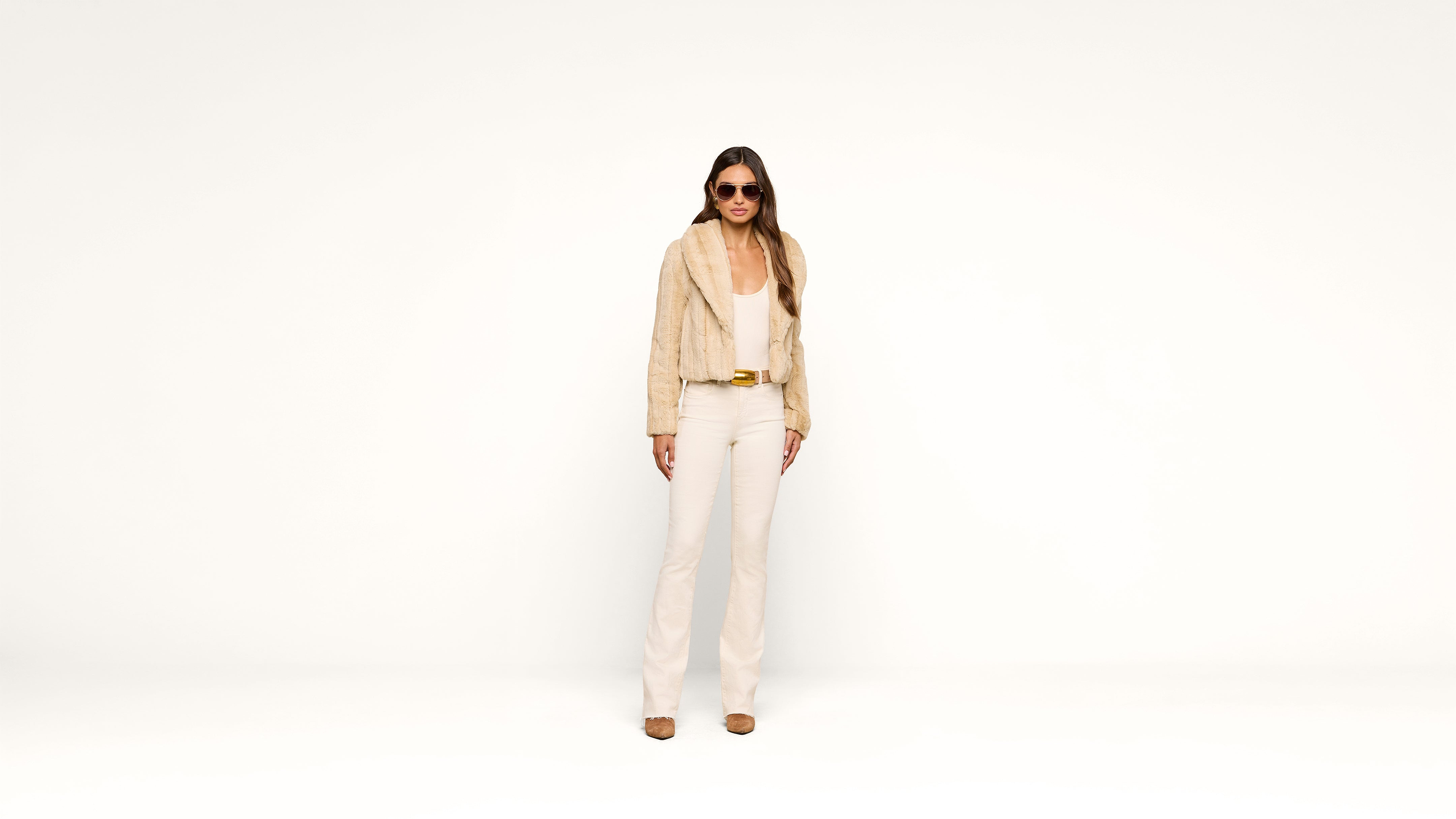 A woman wearing sunglasses, a beige fur jacket, a light top, and cream pants stands against a plain white background, looking forward with a confident pose.