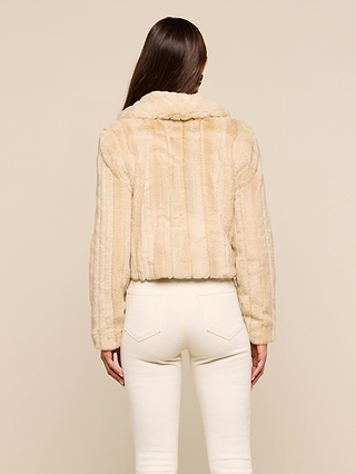A woman stands with her back to the camera against a plain neutral background, wearing the Davy Faux Fur Jacket in beige with a mink look, paired with high-waisted off-white pants.
