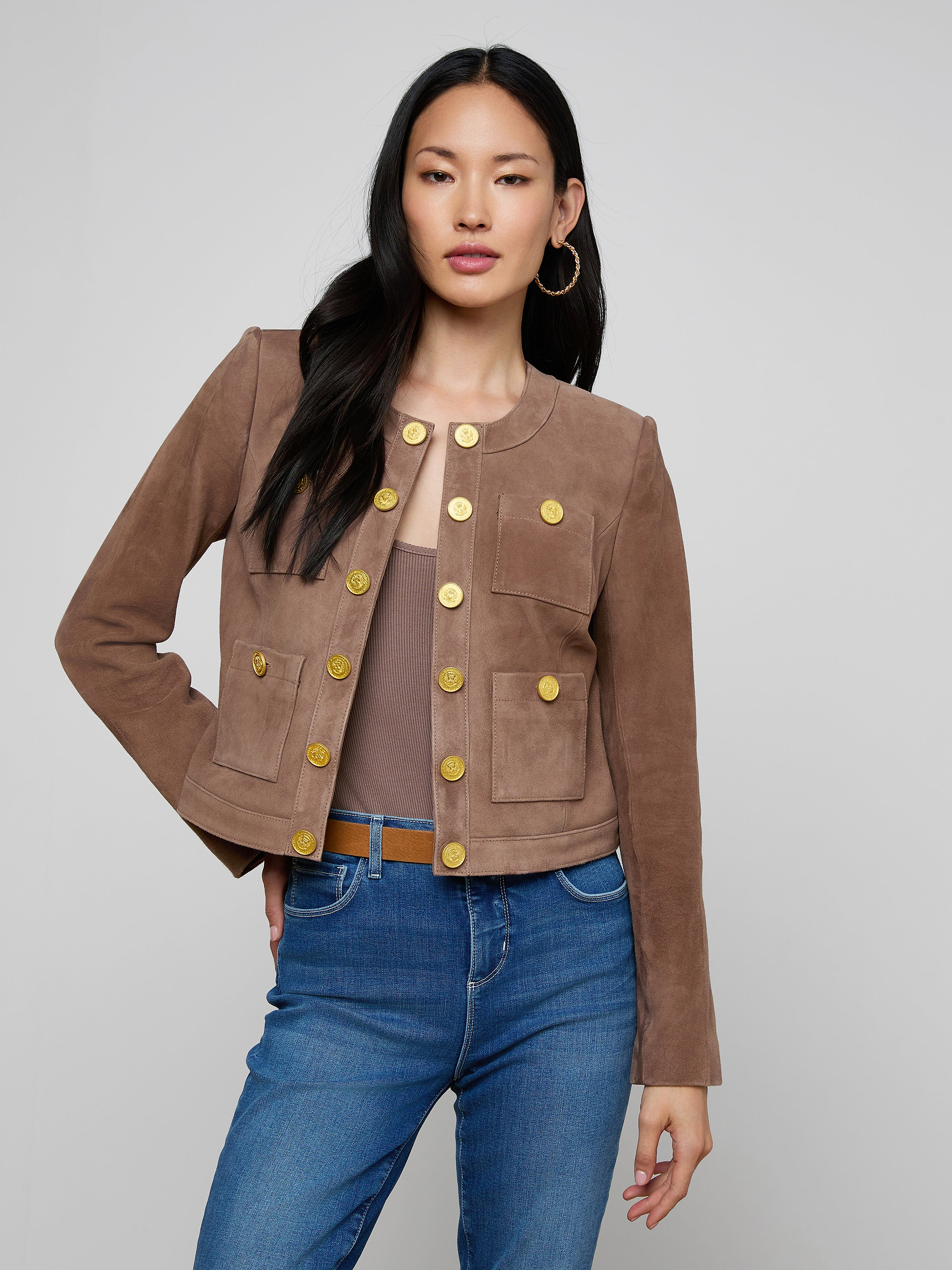 A woman wears the Jayde Collarless Jacket—a brown suede, button-front layering piece—paired with blue jeans as she stands with one hand on her hip against a plain light background. This stylish jacket adds sophistication to her look.