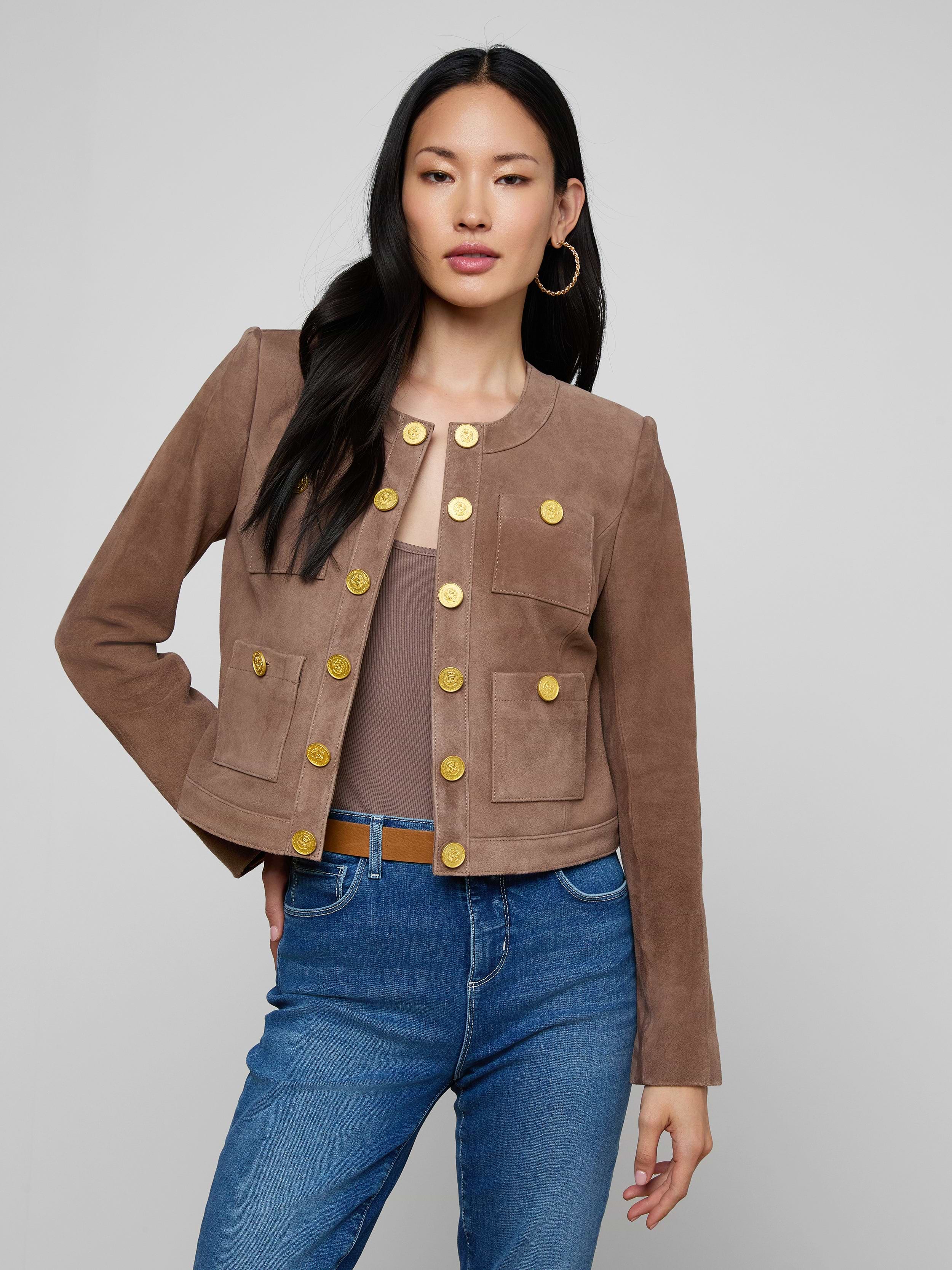 A woman wears the Jayde Collarless Jacket—a brown suede, button-front layering piece—paired with blue jeans as she stands with one hand on her hip against a plain light background. This stylish jacket adds sophistication to her look.