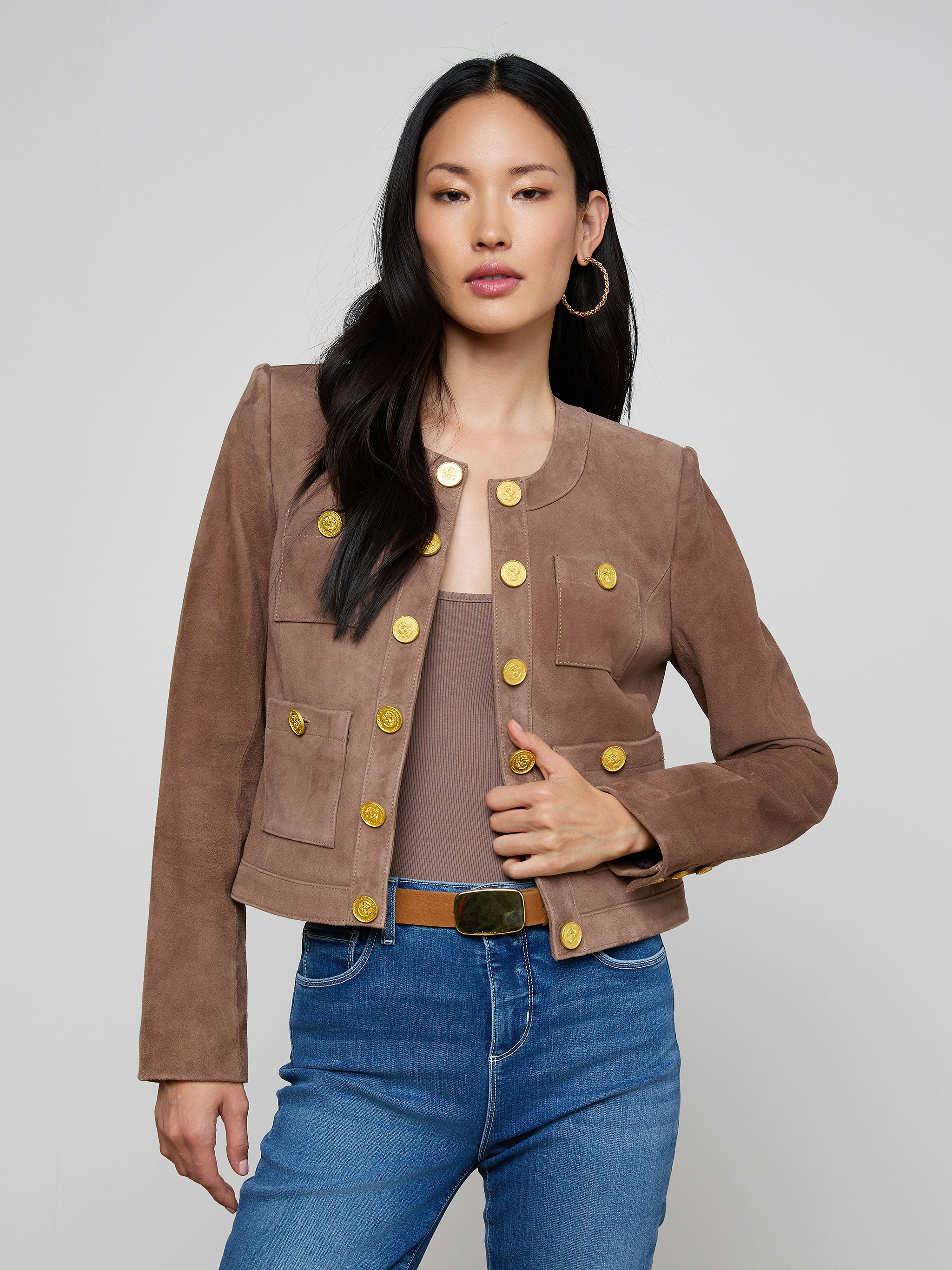 A woman wearing the Jayde Collarless Jacket—a brown suede design with gold buttons—paired with a taupe top and blue jeans, stands with one hand on her hip against a plain light background. This stylish layering piece adds an elegant touch to her look.