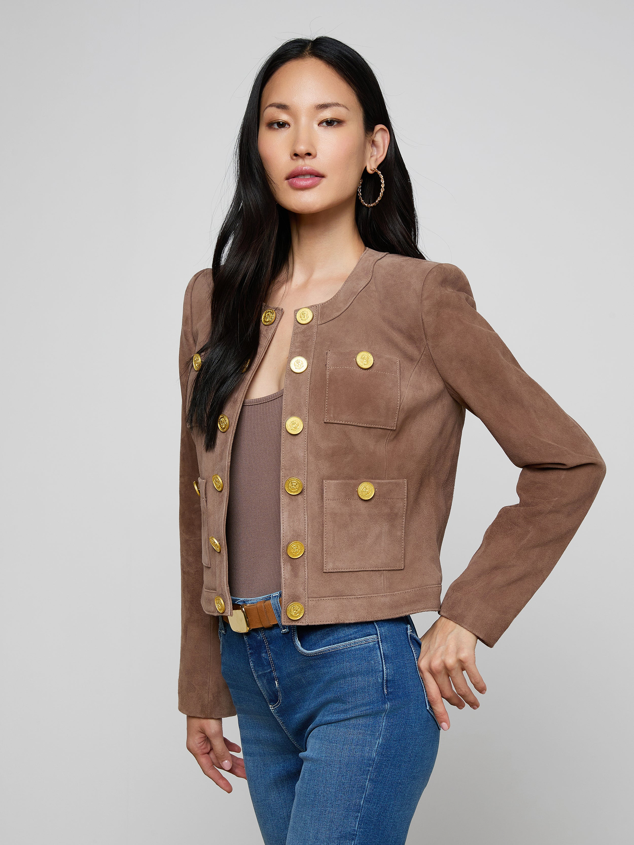 A woman wearing the Jayde Collarless Jacket—a brown suede layering piece with gold buttons—paired with blue jeans, a taupe top, and hoop earrings stands against a plain light background, posing with one hand on her hip. This women’s jacket adds sophisticated style to her outfit.