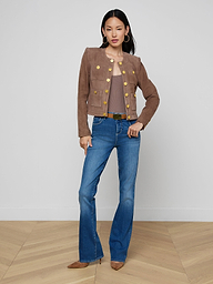 A woman with straight dark hair and a neutral expression stands against a plain white background wearing the Jayde Collarless Jacket with gold buttons, paired with a brown top, blue flared jeans, and brown pointed shoes.