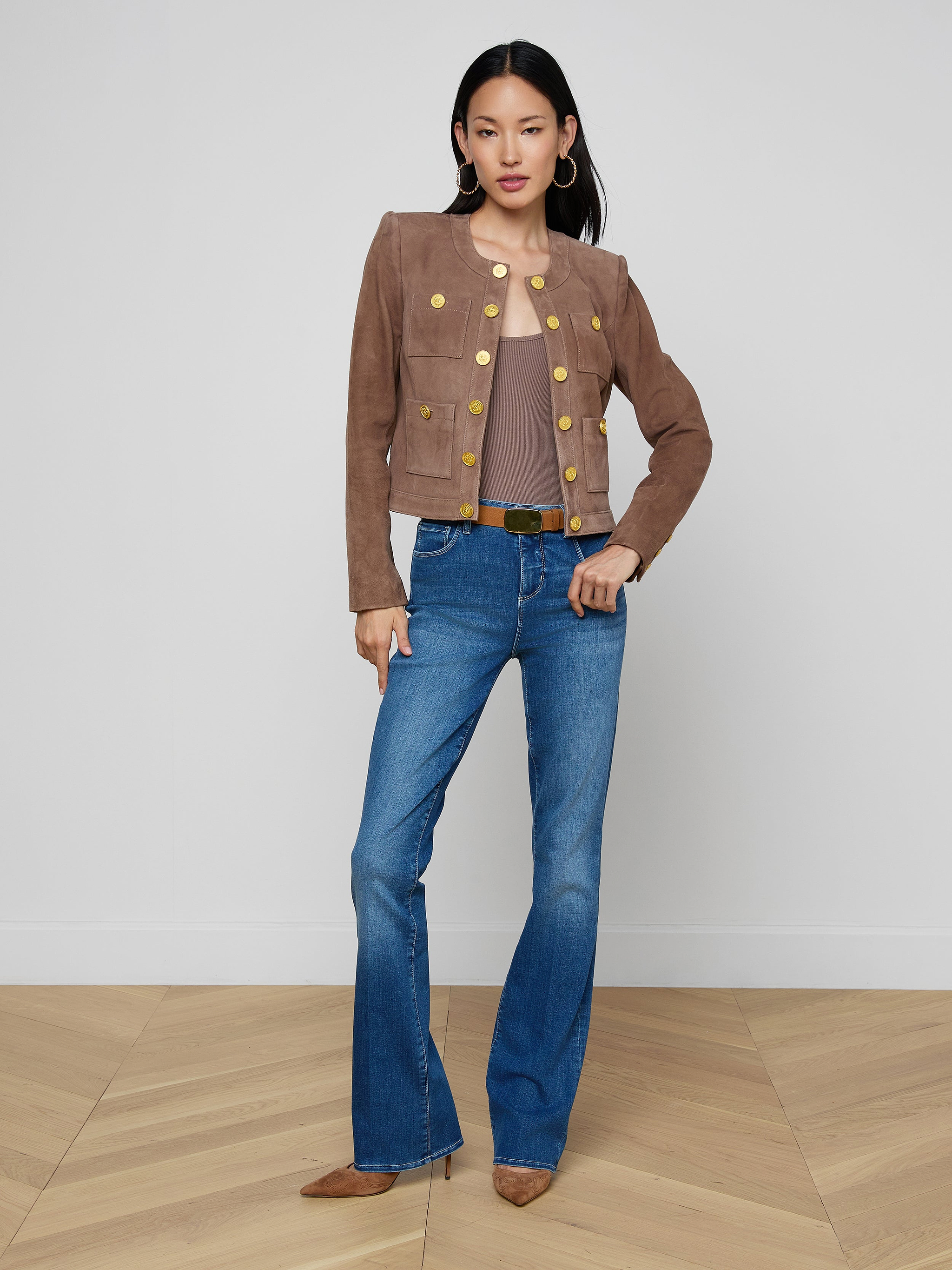 A woman with straight dark hair and a neutral expression stands against a plain white background wearing the Jayde Collarless Jacket with gold buttons, paired with a brown top, blue flared jeans, and brown pointed shoes.